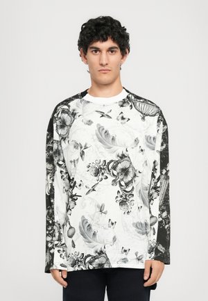 Young man with curly hair wearing a black and white long-sleeve shirt featuring birds, flowers, feathers, and crown patterns.