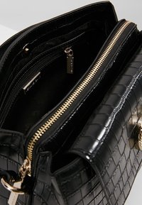 Sac à main noir en simili cuir croco, avec une texture lisse, doté de deux fermetures éclair dorées, de poches intérieures et d'une étiquette métallique de marque.