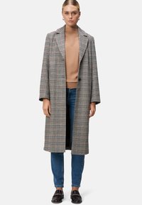 Manteau à motifs à carreaux longs en gris, marron et crème, avec un col cranté, une fermeture à un bouton et des poches latérales, associé à un pull marron.