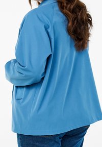 Žena s dlouhými hnědými vlasy, oblečená v volné modré bundě a modrých džínách, otočená mírně do strany s rukama u kapes.