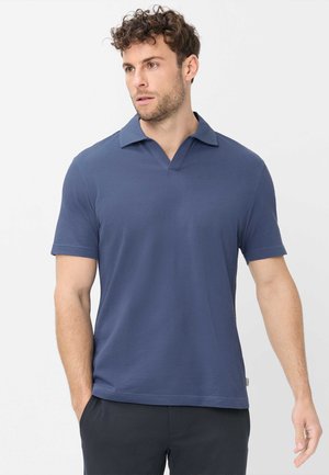 Uomo con capelli ricci che indossa una camicia blu a maniche corte con colletto e pantaloni neri, in piedi con le mani parzialmente nelle tasche, guarda a sinistra.