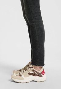 Beige and brown athletic sneakers with a textured upper, lace closure, and a red logo accent. Worn with black jeans featuring frayed hems.