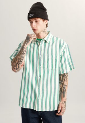 Camisa de manga corta abotonada en blanco con rayas verticales verdes; cuenta con un bolsillo en el pecho, ajuste relajado y textura suave de algodón.