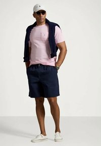 Ανδρική εμφάνιση με ροζ πουκάμισο κοντομάνικο, ναυτικές βερμούδες, λευκά αθλητικά παπούτσια και λευκό καπέλο, με ναυτικό πουλόβερ ριγμένο στους ώμους.