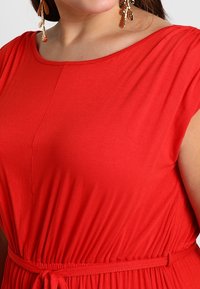 Czerwona sukienka z okrągłym dekoltem, marszczonym materiałem, elastyczną talią i wiązanym paskiem. Teksturowany materiał dodaje subtelnej głębi.