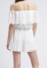 Donna che indossa un completo in pizzo bianco a spalle scoperte con motivi floreali, vita elastica, vista da dietro su uno sfondo semplice.