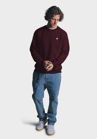 Burgundský svetr s malým žlutým logem slona, v kombinaci s světle modrými džíny a bílými teniskami se hnědými podrážkami.
