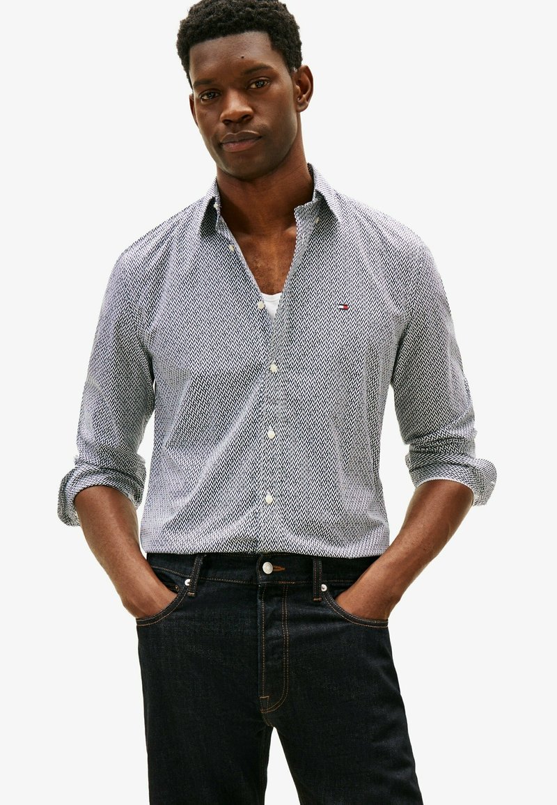 Homme noir aux cheveux courts et bouclés porte une chemise à manches longues à motifs avec les manches retroussées et un jean foncé, les mains dans les poches.