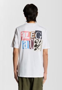 Hvid bomulds T-shirt med et multifarvet grafisk design på ryggen. Teksten inkluderer "Legacy" og "Lyle & Scott" i forskellige skrifttyper og teksturer.