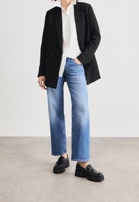 Černý pruhovaný blazer přes bílou košili s knoflíky, v kombinaci se světle modrými širokými kalhotami a černými platformovými mokasíny.