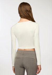 Top corto bianco a maniche lunghe con scollatura tonda, abbinato a pantaloni grigi su misura con tasche posteriori e una texture liscia.