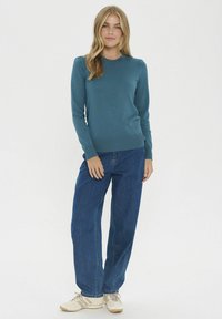 Pull en tricot turquoise avec un col rond, des poignets et un ourlet côtelés, associé à un jean bleu taille haute et des baskets de couleur claire.