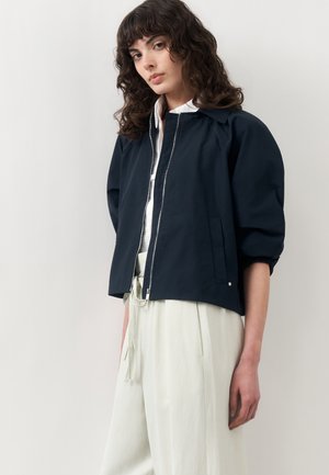 Žena s kučeravými vlasmi oblečená v námornícky modrej bunde na zips cez bielu košeľu a voľné, svetlé nohavice, stojaca pred jednoduchým pozadím.