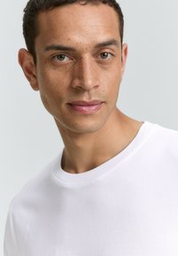 Weißes T-Shirt mit kurzen Ärmeln und rundem Halsausschnitt, getragen von einem männlichen Modell mit kurzen, dunklen Haaren und hellbrauner Haut. Glatte Textur.