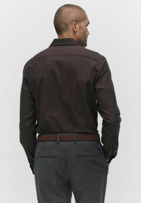 Brun långärmad skjorta med krage, tillverkad av texturerat tyg. Kombinerad med grå byxor och ett brunt bälte, sedd bakifrån.