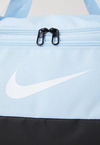 Tote de tela azul claro con un prominente swoosh blanco de Nike, que cuenta con un cierre de cremallera y acentos negros en la parte inferior.