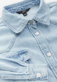 Chaqueta de mezclilla azul claro con botones a presión, cuello puntiagudo y un bolsillo en el pecho, mostrando un detalle en close-up de la tela y las costuras.
