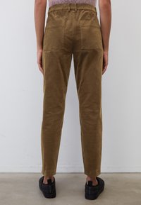 Vrouw in bruine ribfluwelen broek met achterzakken en zwarte instapquilted schoenen, staand op een grijze vloer.