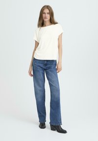 Ung kvinde står iført en hvid kortærmet trøje, løse blå jeans og sorte hæleboots foran en ensfarvet lys baggrund.
