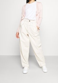 Světlé růžové kardigán překrytý bílým tričkem, doplněný volnými béžovými kalhotami a bílými botami, na šedém pozadí.