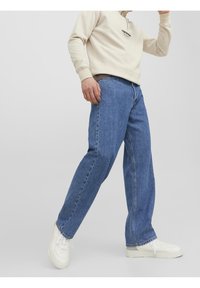 Felpa beige chiaro, jeans in denim blu con una vestibilità comoda e sneakers bianche. I jeans presentano un classico design a cinque tasche e dettagli cuciti.