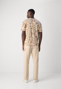 Beige gemustertes Kurzarmhemd mit geometrischen Designs. Kombiniert mit hellbeigen Hosen und weißen Sneakers. Modell von hinten betrachtet.