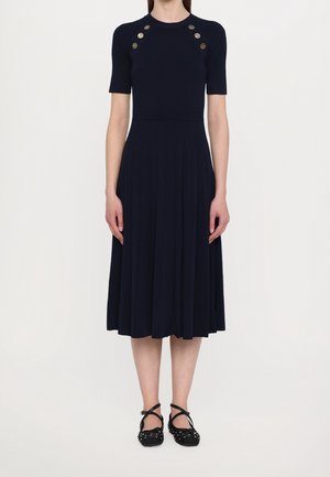 Vestido azul marino acanalado con parte superior ajustada, mangas cortas y falda acampanada. Presenta detalles de botones dorados en los hombros y tela texturizada. Zapatos negros.