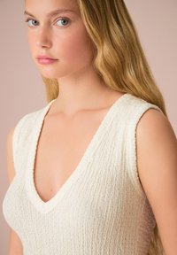 Top senza maniche di colore crema con texture, scollatura a V profonda e design aderente. Il tessuto appare morbido e a coste, mettendo in risalto il décolleté.