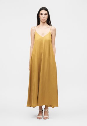 Kvinde med langt mørkt hår iført en sennepsgul ærmeløs maxikjole og hvide strop-sandaler med hæl, stående mod en ensfarvet baggrund.