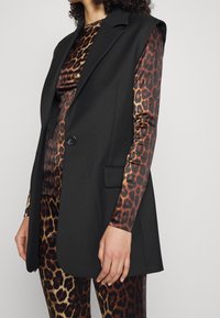 Černý bezrukávový blazer s klopami a jedním knoflíkem, který je spárován s dlouhým rukávem, tričkem s leopardím potiskem v hnědých a zlatých tónech.