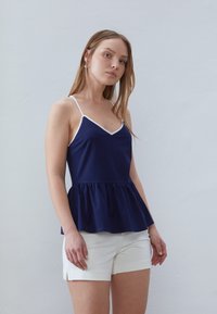Marinblå peplumtop med tunna vita axelband och V-ringning. Tyget verkar slätt, med en utsvängd fåll. Matchas med vita shorts.