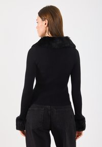 Czarny sweter z ribbowym wzorem, z fałszywym futrzanym kołnierzem i mankietami, dopasowany krój, długie rękawy i teksturowane wykończenie. Styl jest minimalistyczny i monochromatyczny.