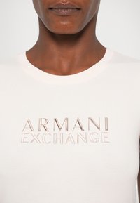 Tricou alb cu guler rotund, având un logo metalic reliefat care spune "ARMANI EXCHANGE" în culori contrastante. Textură de țesătură netedă.