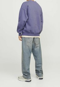 Person in weiten blauen Jeans, weißen Turnschuhen und einem lila Sweatshirt, der mit dem Rücken zu einer einfarbigen, hellen Hintergrund steht.