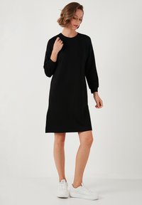 Black long-sleeve dress with a relaxed fit, round neckline, and mid-thigh length; made of soft fabric; paired with white sneakers.