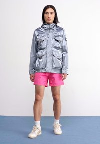 Silver waterproof jacket with multiple front pockets, paired with bright pink shorts. Model wearing white socks and sneakers.