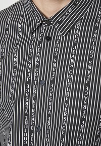 Maglietta a righe verticali bianco e nero con il testo "VERSACE JEANS COUTURE" in motivi ripetuti. Include un colletto e bottoni neri.