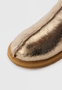 Dessus de chaussure en métal doré avec une finition craquelée, présentant un bout arrondi et une semelle en caoutchouc beige. Matériau texturé, lisse et flexible.
