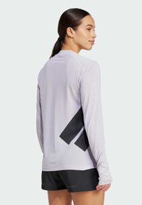 Haut de sport à manches longues en tissu maillé violet clair, présentant un motif géométrique noir dans le dos, avec un col arrondi et une coupe ajustée.