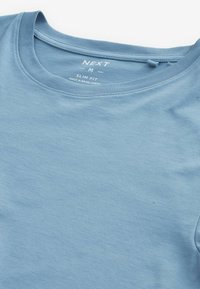 T-shirt en coton slim-fit bleu clair, col rond, taille moyenne, fabriqué au Bangladesh, marque "NEXT" visible à l'intérieur du col.