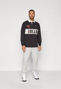 Črna polo majica z dolgimi rokavi in okroglim izrezom, bež črtami, napis "BULLS" in rdečim logotipom bik. V paru s sivimi cargo hlačami in belimi športnimi copati.