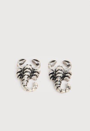 Paire de petites boucles d'oreilles en métal argenté en forme de scorpion avec des pinces relevées et des queues enroulées sur un fond blanc uni.