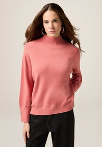 Maglione rosa a maglia con un alto colletto a coste e maniche lunghe, caratterizzato da orli a coste e una leggera trama. Abbinato a pantaloni neri.