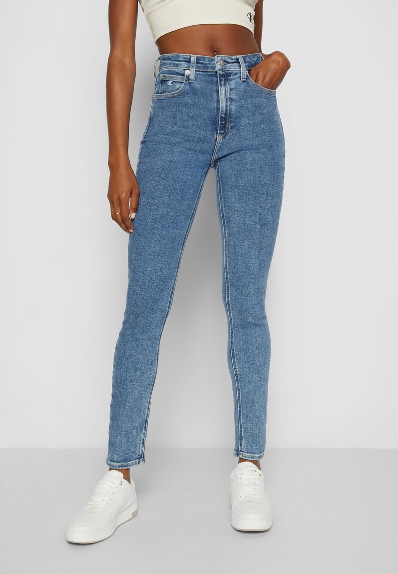 Person iført højtaljede blå skinny jeans med hånden i lommen, hvid cropped top og hvide sneakers, stående foran en ensfarvet baggrund.