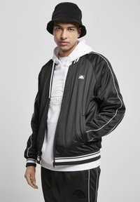 Giacca bomber nera con strisce bianche, abbinata a un cappuccio bianco. Il modello indossa un cappello a secchiello in corduroy nero. Texture liscia, stile casual.