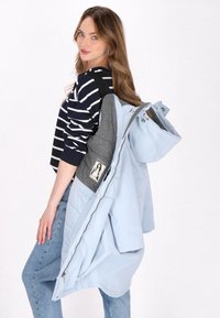Jachetă pufosă de culoare bleu deschis, cu glugă și accente gri, având un buzunar cu un grafic. Un top cu dungi și blugi bleu deschis completează ținuta.