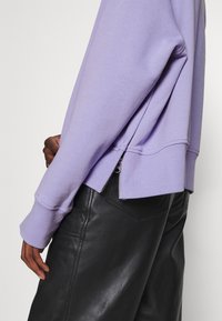 Sudadera lila de corte relajado, con un detalle de cremallera en el dobladillo. Combinada con pantalones de cuero negros, mostrando una textura suave.