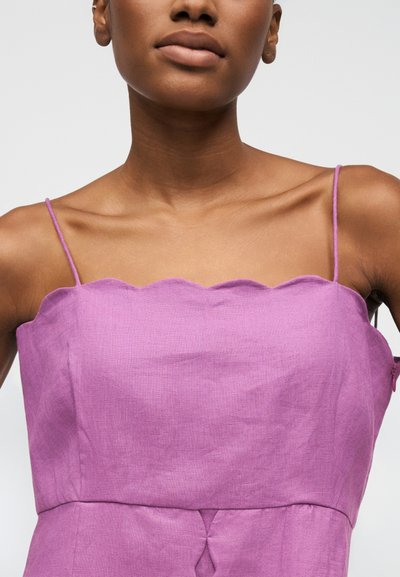 Gros plan sur une personne portant une robe violette sans manches avec un décolleté festonné et de fines bretelles, sur un fond uni.