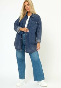 Camicia di jeans blu scuro con chiusura a bottoni, due tasche sul petto, vestibilità oversized e polsini a contrasto, abbinata a jeans a gamba larga blu chiaro e sneakers bianche.