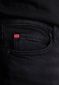 Jean en denim noir avec une poche arrière arborant une petite étiquette rouge "HUGO", texture lisse et détail de couture subtil.